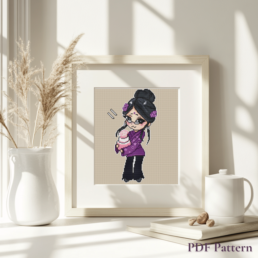 Framed Aunt Karma cross stitch pattern displayed in a bright cozy interior with dried pampas grass, a ceramic vase, and a cup, showing how the finished embroidery looks as home decor.